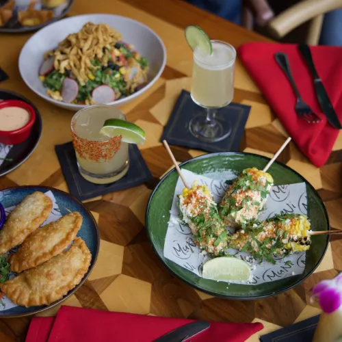 Overhead spread of MuNi KC shareables: street corn, empanadas, egg rolls, salad and cocktails on a wood table
