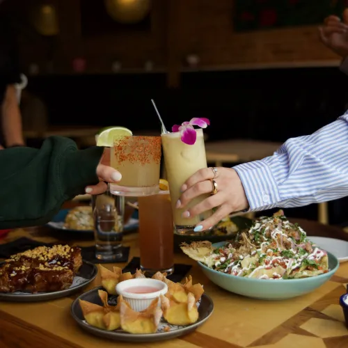 Two guests toasting tropical cocktails over a shared plate of nachos at MuNi KC